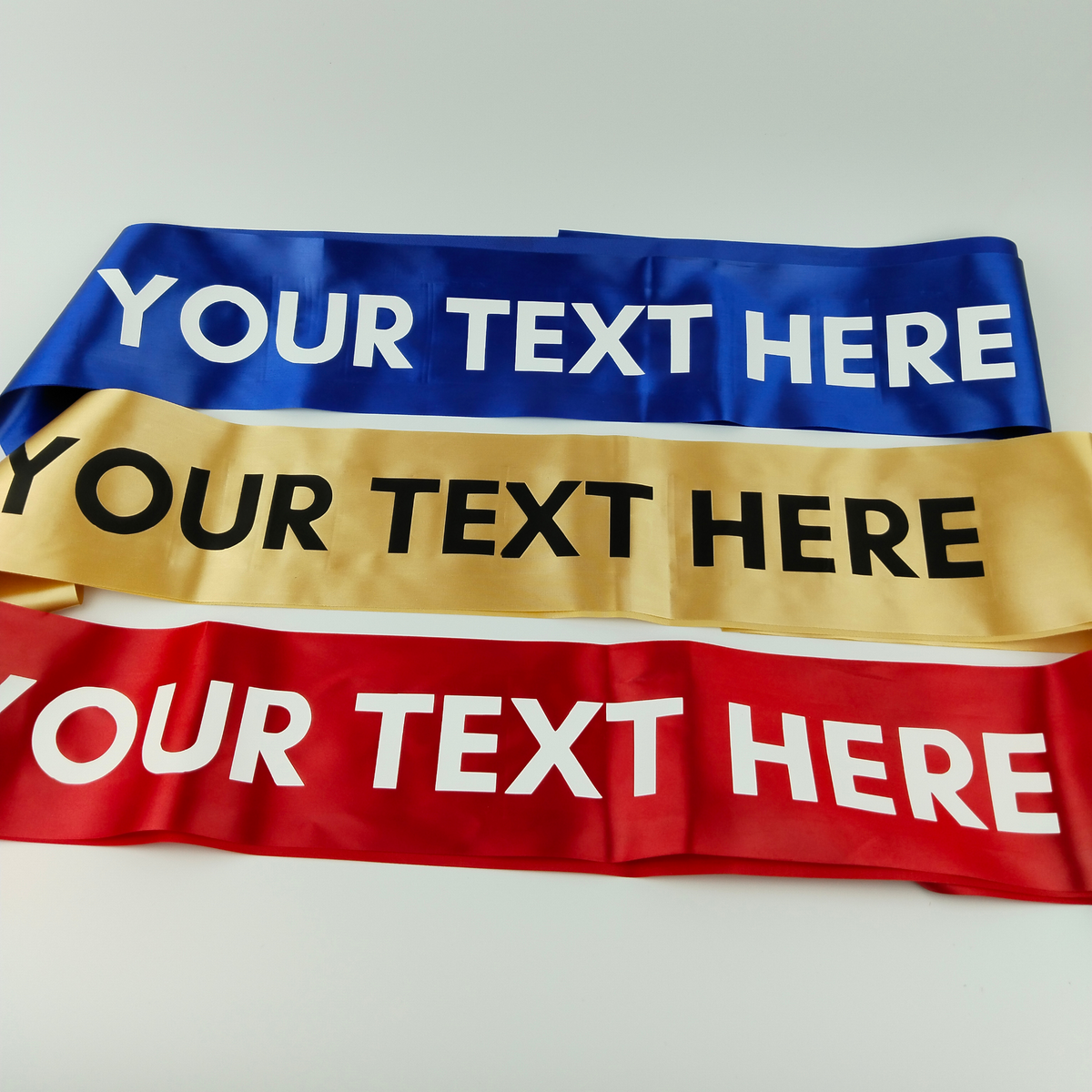 Three satin sashes displayed horizontally in red, gold, and blue colors with text areas for custom personalization.